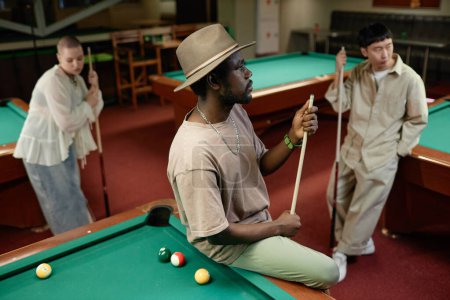 Téléchargez les photos : Portrait d'un homme adulte afro-américain portant un chapeau avec un groupe multiethnique de personnes dans un club de billard - en image libre de droit