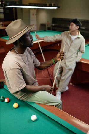 Photo for Vertical portrait of Black adult man wearing hat posing on pool table in bar - Royalty Free Image