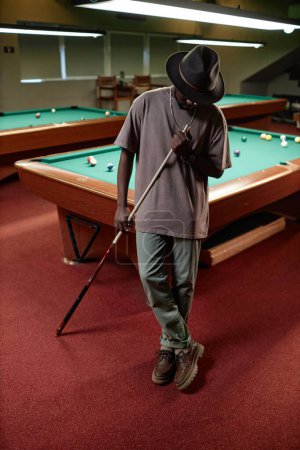 Téléchargez les photos : Portrait vertical pleine longueur d'un homme adulte noir portant un chapeau posé par une table de billard - en image libre de droit
