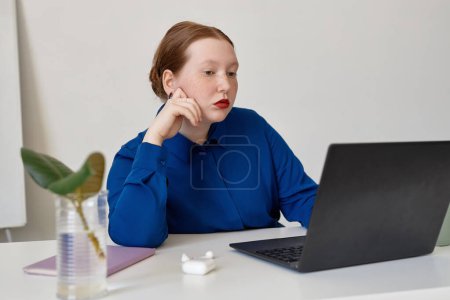 Portrait of creative red haired woman thinking on design project and using laptop in office, copy space