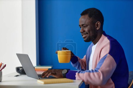 Téléchargez les photos : Vue latérale vibrante portrait de l'homme noir en utilisant un ordinateur portable sur le lieu de travail dans le bureau avec horloge sur le mur bleu, espace de copie - en image libre de droit