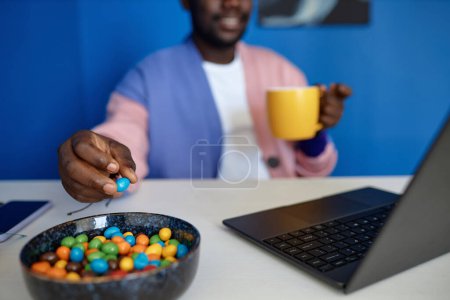 Foto de Primer plano del hombre negro disfrutando de bocadillos de caramelo y café mientras usa la computadora portátil en la oficina vibrante, espacio para copiar - Imagen libre de derechos