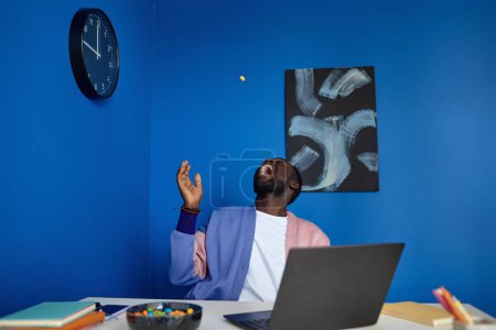 Photo for Portrait of creative black man having fun in colorful office interior and throwing candy in air, copy space - Royalty Free Image
