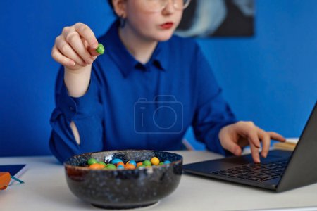Foto de Primer plano de la mujer joven disfrutando de bocadillos de caramelo mientras usa el ordenador portátil en la oficina vibrante, espacio para copiar - Imagen libre de derechos