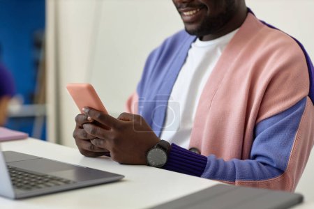 Téléchargez les photos : Gros plan d'un homme noir souriant tenant un smartphone sur le lieu de travail dans un bureau coloré, espace de copie - en image libre de droit