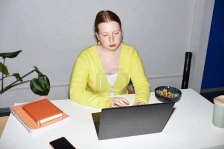 Téléchargez les photos : Portrait propre de jeune femme rousse utilisant un ordinateur portable sur le lieu de travail tourné avec flash - en image libre de droit