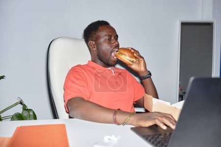 Téléchargez les photos : Portrait latéral de l'homme noir mangeant un hamburger sur le lieu de travail et utilisant un ordinateur portable, tourné avec flash - en image libre de droit