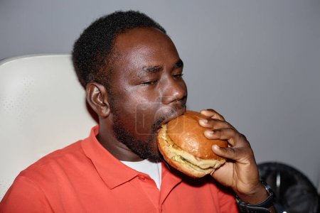Foto de Mínimo retrato de vista lateral del hombre negro comiendo hamburguesa en el interior disparado con flash, espacio de copia - Imagen libre de derechos