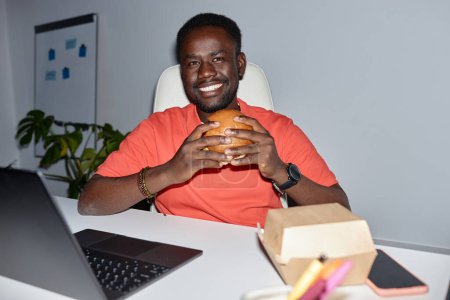 Téléchargez les photos : Vue de face portrait d'un homme noir souriant mangeant un hamburger sur le lieu de travail tourné à l'intérieur avec flash - en image libre de droit