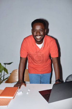 Téléchargez les photos : Portrait vertical de l'homme noir souriant regardant la caméra sur le lieu de travail et penché sur le bureau, tourné avec flash - en image libre de droit