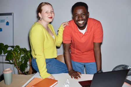Téléchargez les photos : Vue de face portrait de deux personnes souriantes regardant la caméra sur le lieu de travail et penchées sur le bureau, prises de vue avec flash - en image libre de droit