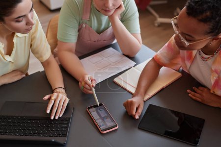 Photo for Close up of three young women discussing UI UX design project at meeting table in office and pointing at smartphone screen - Royalty Free Image