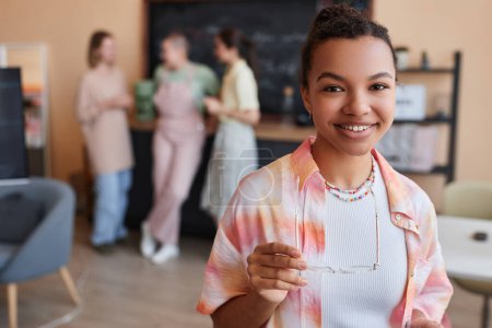 Portrait of young gen Z black woman smiling at camera in office setting and wearing colorful casual clothes, copy space