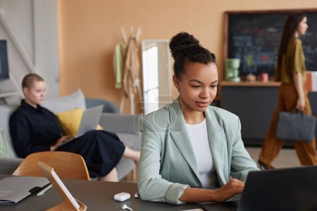 Portrait of young black businesswoman using laptop in modern office with all female team, copy space