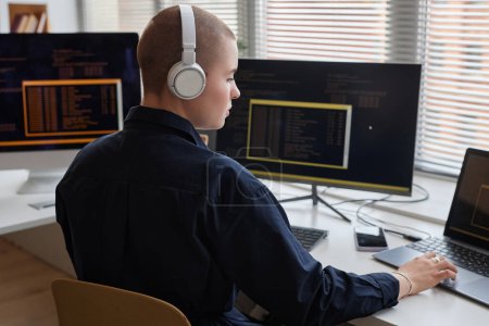 Retrato de vista lateral de una joven calva que usa auriculares y computadora mientras escribe código y trabaja en TI