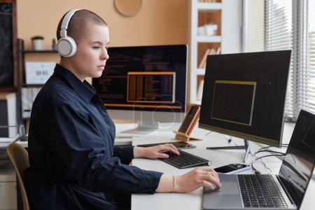 Portrait of female programmer wearing headphones and using computer while writing code and working in IT