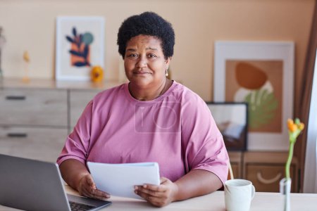 Aged African American businesswoman in casual attire looking at camera while sitting by desk in front of laptop and working at home office