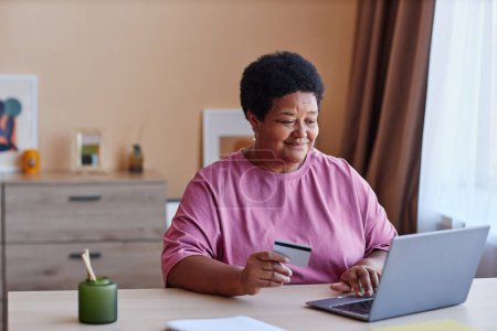 Téléchargez les photos : Femme âgée assise par bureau devant un ordinateur portable dans le salon et saisissant les données de sa carte de crédit lors de la commande de produits dans la boutique en ligne - en image libre de droit