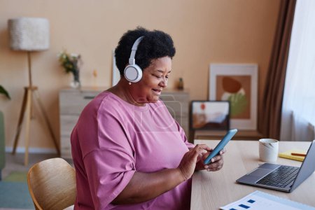 Vue latérale d'une femme mûre souriante dans un casque et un t-shirt rose regardant à travers la playlist dans un smartphone tout en choisissant quelque chose à écouter