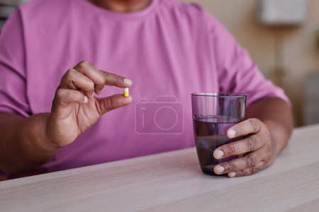 Close-up of sick African American senior woman holding small pill and glass of water while sitting by table and going to take medicaments