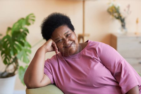 Téléchargez les photos : Happy African American senior femme en t-shirt rose assis dans le fauteuil devant la caméra et vous regarder avec le sourire dans l'environnement de la maison - en image libre de droit