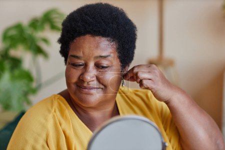 Femme mûre souriante essayant de petites boucles d'oreilles en perles tout en se tenant devant la caméra et en regardant dans le miroir pendant la préparation de la date