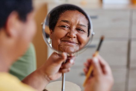 Focus on reflection of happy mature African American woman face in round mirror during process of applying makeup as part of daily routine