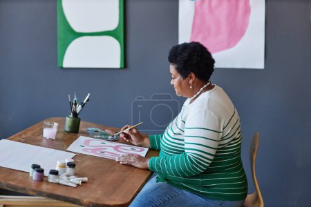 Photo for Side view of retired woman with paintbrush creating new artwork or masterpiece with gouache of various colors by workplace in studio - Royalty Free Image