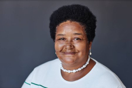 Foto de Retrato de mujer afroamericana envejecida en jersey blanco, collar de perlas y pendientes mirando a la cámara con sonrisa contra la pared gris en el estudio - Imagen libre de derechos