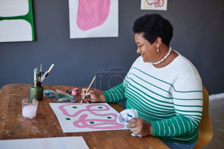 Foto de Feliz artesana senior con pincel y pintura gouache en hoja de papel mientras se sienta a la mesa en el estudio de artes y artesanía en el ocio - Imagen libre de derechos