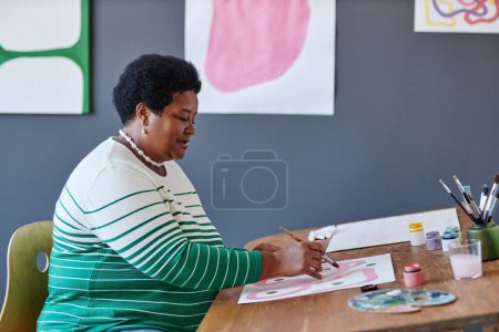 Seitenansicht der reifen Rentnerin mit Pinselmalerei auf Papierblatt mit rosa Gouache, während sie am Schreibtisch im Atelier der Künste sitzt