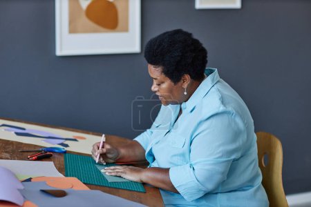 Foto de Vista lateral de la anciana artesana afroamericana que trabaja sobre nuevas obras de arte mientras está sentada en el lugar de trabajo y dibuja la línea en un pedazo de papel - Imagen libre de derechos