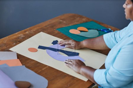 Foto de Manos de artesana afroamericana madura creando una nueva composición de papel de varios colores mientras está sentada junto a un escritorio de madera en el estudio - Imagen libre de derechos