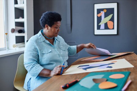 Photo for Aged African American female designer working with colorful paper while creating new composition on white sheet by workplace in studio - Royalty Free Image