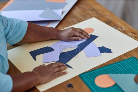 Hände eines reifen afroamerikanischen kreativen Designers, der im Atelier eine neue Papierkomposition auf weißem Blatt auf einem Holztisch kreiert