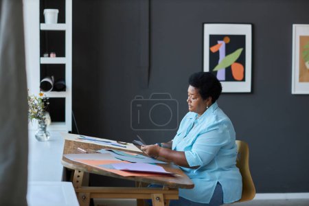 Téléchargez les photos : Vue latérale de créatif senior femme noire en casual papier de coupe avec des ciseaux tout en travaillant sur la création de nouveau chef-d'œuvre en studio - en image libre de droit