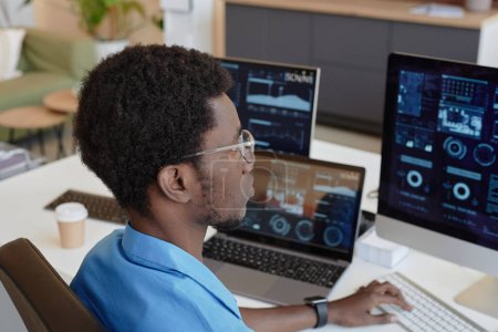 Téléchargez les photos : Jeune homme noir portant des lunettes regardant des graphiques et des graphiques sur des écrans d'ordinateur assis par lieu de travail dans un bureau et analysant des données - en image libre de droit