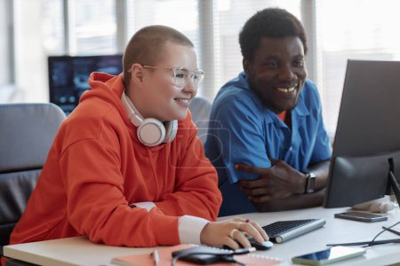 Téléchargez les photos : Deux jeunes ingénieurs interculturels heureux en cybersécurité assis sur le lieu de travail devant un écran d'ordinateur et regardant à travers les données - en image libre de droit