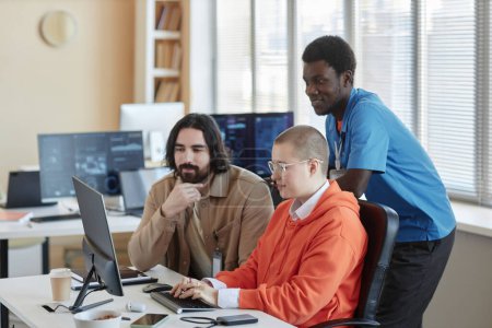 Junge Geschäftsfrau tippt auf Computertastatur, während ihre beiden interkulturellen männlichen Kollegen beim Arbeitstreffen auf den Bildschirm schauen