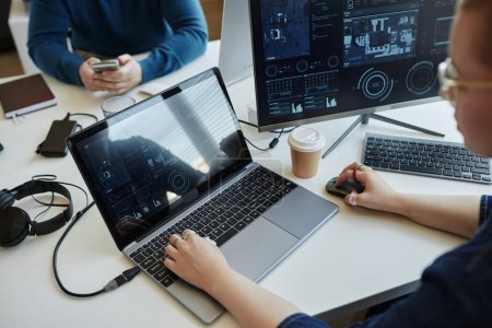 Junge Analytikerin oder IT-Managerin mit Laptop am Arbeitsplatz vor Computermonitor mit grafischen Daten