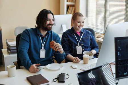 Zwei glückliche junge Büromanager bei Snacks in der Mittagspause, während sie vor dem Computerbildschirm sitzen und neugierige Online-Videos ansehen