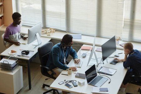 Oberhalb des Winkels mehrerer interkultureller IT-Ingenieure, die im Büro vor Computerbildschirmen sitzen und Online-Daten oder Statistiken analysieren