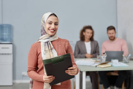 Junge lächelnde multiethnische Unternehmensführerin mit Folder, die Sie gegen männliche und weibliche Kollegen ansieht, die Ihre Präsentation vorbereiten