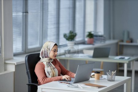 Téléchargez les photos : Jeune musulmane sérieuse ceo dactylographier sur clavier d'ordinateur portable tout en étant assis par le lieu de travail dans un bureau ouvert et l'analyse de l'information en ligne - en image libre de droit