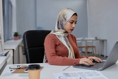 Junge ernstzunehmende muslimische Spezialistin für Kopftuch arbeitet an neuem Projekt oder analysiert Online-Daten beim Tippen auf Laptop-Tastatur