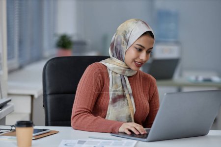 Junge lächelnde muslimische Geschäftsfrau mit Kopftuch sitzt am Arbeitsplatz vor dem Laptop und schaut sich Online-Informationen auf dem Bildschirm an
