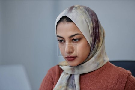 Photo for Young pretty Muslim female employee in headscarf looking at laptop or other gadget screen while sitting in front of camera during network - Royalty Free Image