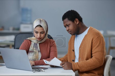Junger afroamerikanischer Geschäftsmann mit Dokument schaut auf Laptop-Bildschirm, während er neben Kollegin sitzt, die Präsentation macht