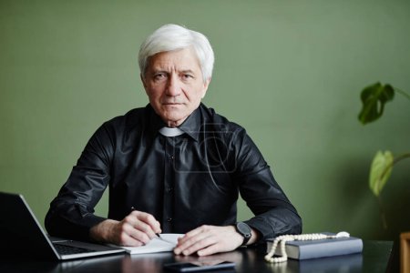 Photo for Front view portrait of white haired senior priest working at desk and looking at camera in office against green wall, copy space - Royalty Free Image