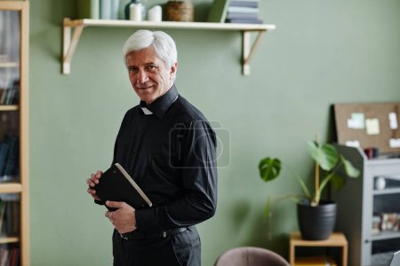 Téléchargez les photos : Portrait taille haute du prêtre aîné souriant regardant la caméra et tenant la Bible tout en restant au bureau contre un mur vert, copiez l'espace - en image libre de droit
